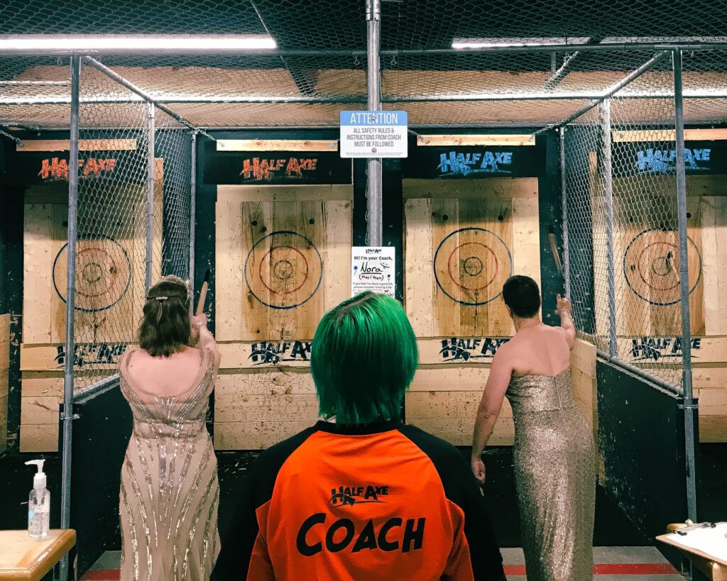 Two women in fancy dresses facing away from camera throwing hatchets at targets. In the middle of the picture is a Coach (on their shirt) watching them throw. 